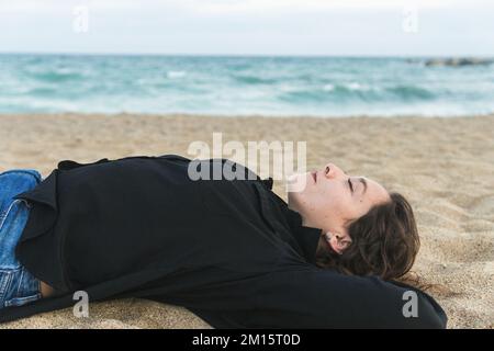 Seitenansicht einer glücklichen jungen Frau mit lockigem, lächelndem Haar und während sie am Sandstrand liegt Stockfoto