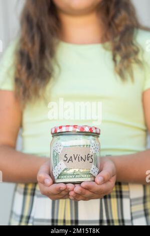 Schneiden Sie ein Mädchen mit langen Haaren, das einen Glasbehälter hält, mit Spareinschrift und Geld im selektiven Fokus Stockfoto