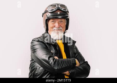 Porträt eines lächelnden, gealterten, bärtigen männlichen Bikers in Lederjacke und Motorradhelm, der mit gekreuzten Armen auf weißem Hintergrund in die Kamera schaut Stockfoto