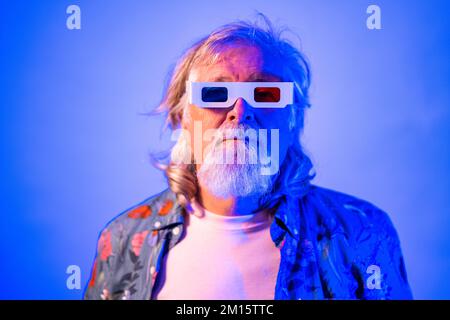 Verblüffter alter Mann mit dreidimensionaler Brille, der mit offenem Mund auf blauem Hintergrund in die Kamera schaut Stockfoto