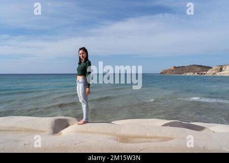 Eine ethnische Frau mit seitlichem Blick, die auf einer felsigen Klippe in der Nähe von klapperndem blauem Wasser gegen den bewölkten Himmel während der Reise durch Italien steht Stockfoto