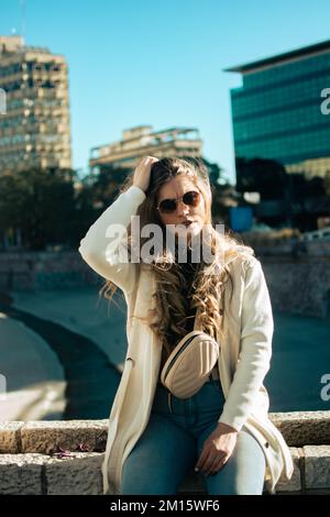 Ein vertikales Bild einer jungen Frau in einer leichten Jacke mit Sonnenbrille, die auf einer niedrigen Steinwand auf der Straße sitzt Stockfoto