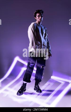 Der ganze Körper eines jungen männlichen Models in Zwangsjacke auf lila Hintergrund steht vor der Kamera vor Neonstrahlen Stockfoto