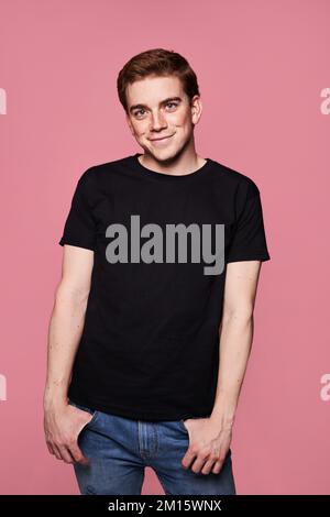 Positiver junger Mann in schwarzem T-Shirt, der Hände in Jeans-Taschen hält und mit einem Lächeln vor pinkfarbenem Hintergrund in die Kamera schaut Stockfoto
