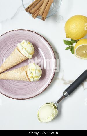 Blick von oben auf appetitliches Eis in Waffelkornets mit der Schale auf dem Keramikteller neben frischen Zitronen und einer Schüssel mit Zimtstangen Stockfoto