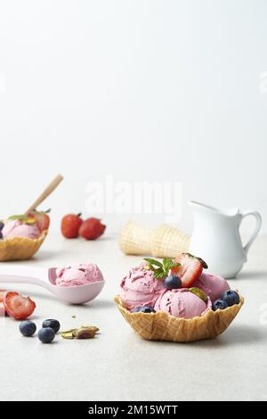 Leckere Beereneisschaufeln auf knusprigen Waffelschüsseln, dekoriert mit frischen reifen Erdbeeren, Blaubeeren, Pistazien und Minzblättern auf weißem Hintergrund Stockfoto