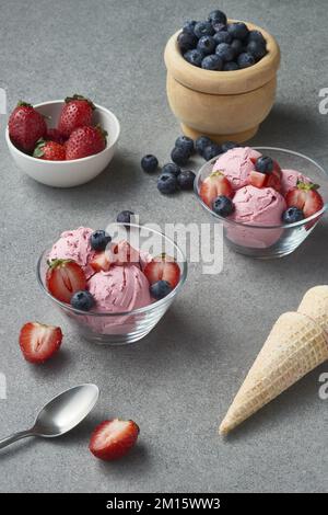 Von oben mit Glasschüsseln mit köstlichen hausgemachten Eislöffeln, dekoriert mit frischen Erdbeeren und Blaubeeren und auf grauem Tisch mit Waffeln Stockfoto