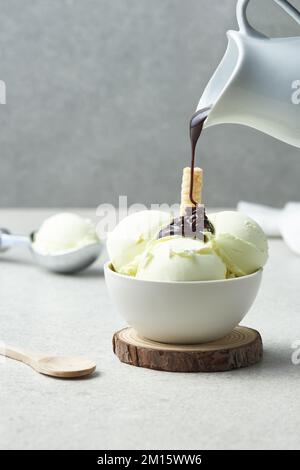 Eine anonyme Person gießt Schokoladenganache aus dem Krug auf köstliche Vanilleeis in einer Keramikschüssel, serviert auf einem kleinen Holzbrett auf dem Tisch Stockfoto