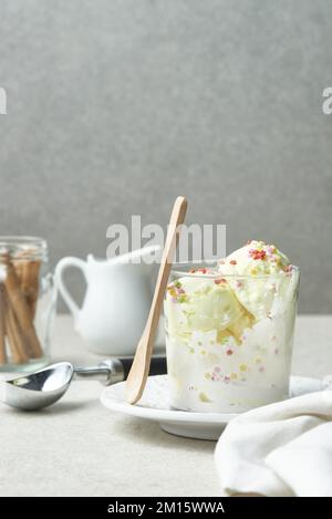 Schaufeln mit köstlichem Vanilleeis mit bunten süßen Streuseln, serviert in einem Glasbecher auf dem Tisch mit einem Holzlöffel aus Zimtstangen und einem Krug Stockfoto