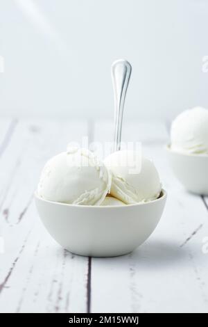 Kübel Vanilleeis in weißen Keramikschüsseln mit Löffeln neben Waffelkonen auf einem weißen Tisch Stockfoto
