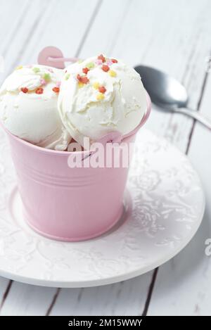 Das Vanilleeis mit hohem Appetit wird in einem kleinen rosa Metalleimer serviert und mit süßen Sternenstreuseln auf einem weißen Holztisch dekoriert Stockfoto