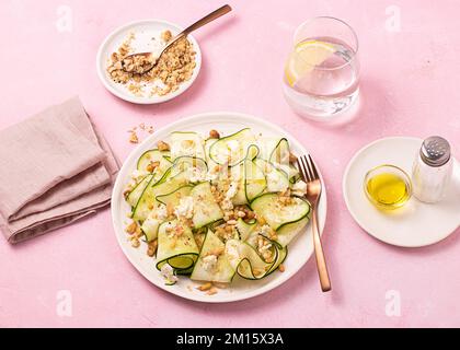 Von oben frischer Salat aus dünnen Scheiben reifer Zucchini, Fetakäse und Pinienkerne auf weißem Teller auf rosa Tischhintergrund Stockfoto