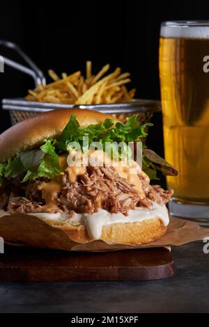 Leckerer Burger mit Schweinefleisch mit Barbecue-Sauce, Cheddar-Käse und Salat, serviert mit Pommes Frites und einem Glas kaltem Bier auf Holzbrett Stockfoto