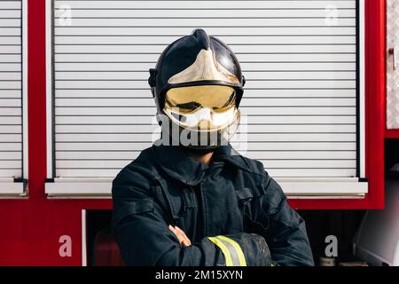 Nicht wiedererkennbarer selbstsicherer anonymer Feuerwehrmann in Schutzuniform und Helm, der die Arme kreuzt, während er gegen das Garagentor steht Stockfoto