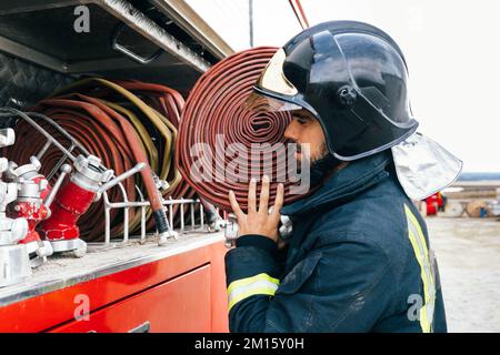 Seitenansicht des hispanischen Feuerwehrmanns in Uniform und Helm mit Schlauch auf der Schulter, der tagsüber in die Nähe des Feuerwehrwagens blickt Stockfoto