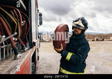 Seitenansicht des hispanischen Feuerwehrmanns in Uniform und Helm mit Schlauch auf der Schulter, der tagsüber in die Nähe des Feuerwehrwagens blickt Stockfoto