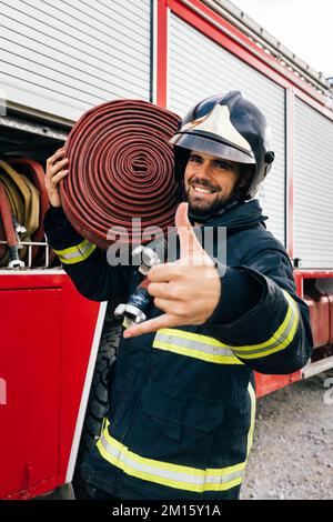 Happy Hispanic Feuerwehrmann in Uniform und Helm trägt Schlauch auf der Schulter und Gesten shaka Schild, während er tagsüber neben dem Feuerwehrauto stand Stockfoto