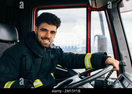 Fröhlicher hispanischer Feuerwehrmann in Schutzkleidung, der beim Führen eines Feuerwehrwagens lächelt und in die Kamera schaut Stockfoto