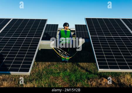 Der ganze Körper eines ruhigen bärtigen Installateurs im Helm blickt weg, während er in der Nähe von Photovoltaikzellen auf dem Hintergrund des blauen Himmels bei Tageslicht steht Stockfoto