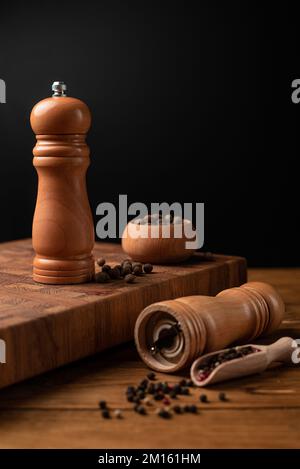 Pfeffermühle und ganzer Pfeffer in Holzschaufel auf schwarzem Tisch. Stockfoto