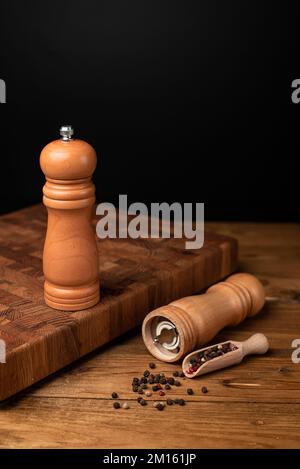 Pfeffermühle und ganzer Pfeffer in Holzschaufel auf schwarzem Tisch. Stockfoto