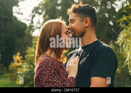 Ein fröhlicher junger Mann küsst seine Frau auf die Stirn, während er an einem sonnigen Tag im Park umarmt. Stockfoto