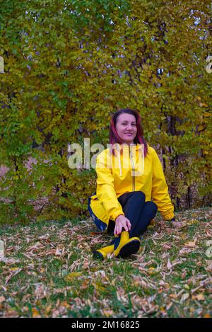 Ein Mädchen sitzt auf dem Rasen in einem gelben Regenmantel und Stiefeln Stockfoto