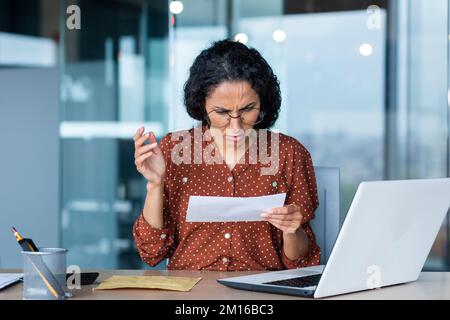 Eine junge lateinamerikanische Frau, Sekretärin, Geschäftsführerin, Buchhalterin, hält beunruhigend einen Brief, ein Dokument, ein Konto. Sie sitzt verwirrt im Büro am Schreibtisch. Stockfoto