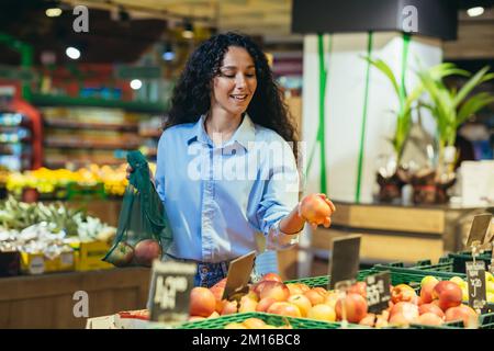 Lächelnde, wunderschöne lateinamerikanische Geschäftsfrau wählt und kauft frisches Obst im Supermarkt für Restaurant, Zuhause. Er hält Äpfel und einen Öko-Sack. Stockfoto