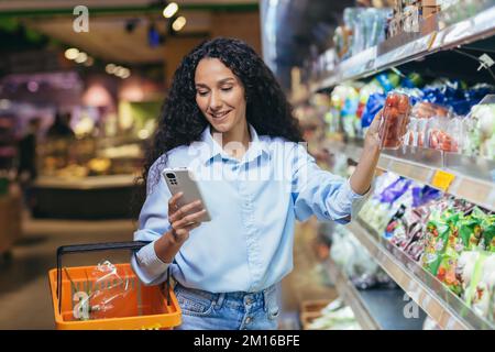 Eine lateinamerikanische Frau steht in einem Supermarkt mit einem Korb neben dem Kühlschrank und Gemüse. Er hält das Telefon in der Hand, liest die Liste, die Zusammensetzung der Produkte, wählt Kirschtomaten. Stockfoto