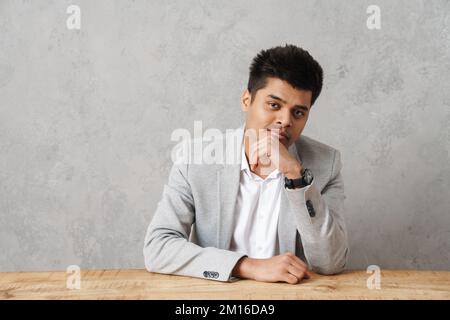 Junger brauner indischer Geschäftsmann, der lächelt und an einem Tisch im Büro über einer grauen Wand sitzt Stockfoto