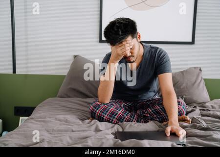 Der junge, müde indische Mann schloss die Augen mit der Hand und hielt den Laptop zu Hause auf dem Bett Stockfoto