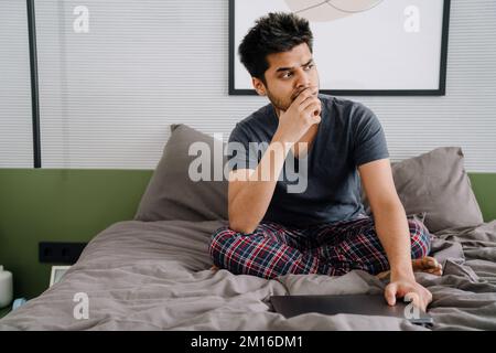 Ein junger, indischer, aufmerksamer Mann, der das Kinn mit der Hand berührt und seinen Laptop geschlossen hält. Er sieht nach rechts aus und sitzt zu Hause auf dem Bett Stockfoto
