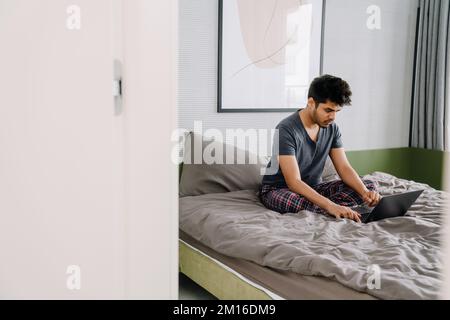 Junger, gutaussehender indianer, der zu Hause auf dem Bett sitzt und an einem Laptop arbeitet, in einem gemütlichen Schlafzimmer Stockfoto