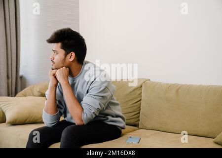 Junger, nachdenklicher indianer, der auf dem Sofa sitzt und zu Hause denkt Stockfoto