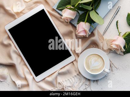 Minimalistischer Arbeitsplatz mit digitalem Tablet-leerem Bildschirm-Mockup, gemütlichem Schal, einer Tasse Kaffee und Rosenblumen auf betontem Hintergrund. Draufsicht, flach liegend Stockfoto