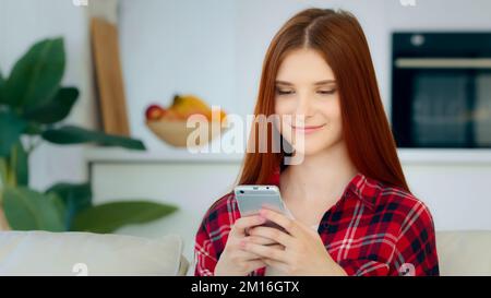 Lächelnd rothaarige kaukasische Mädchen Modell Frau sitzt im Küchenraum hält moderne Gadget Smartphone schaut auf mobile Display liest sms-Nachrichten aus Stockfoto