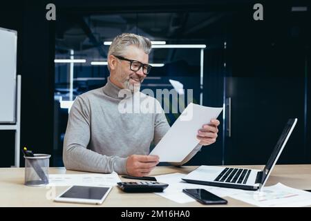 Erfolgreicher Geschäftsmann und Investor, der im Büro an Papierarbeiten arbeitet, reifer, grauhaariger männlicher Boss, der mit Verträgen arbeitet und Finanziers, die Laptops bei der Arbeit verwenden. Stockfoto