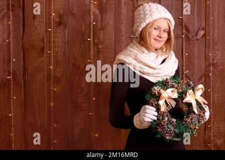 Mädchen mit weihnachtsbaum-Kranz dekorieren zu Hause Holzwände. Junge Frau in Winterstrickkleidung auf braunem Holzhintergrund Stockfoto