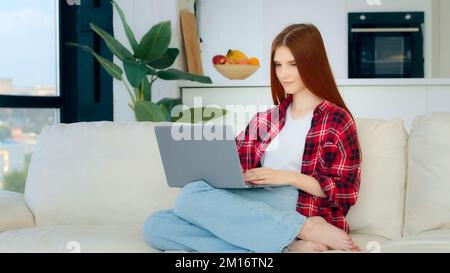 Junge Studentin kaukasischen Freiberufler Hausfrau in legerer Kleidung karierten kurze Geschäftsfrau Mädchen sitzt auf Sofa Suche Stellenangebot im Internet-Check-E-Mail Stockfoto