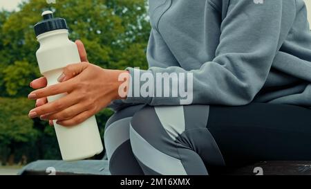 Nahaufnahme unbekannter Athlet gesunde Frau Läuferin trägt Sportanzug Sitzen Sie auf der Bank im Park Ruhe nach dem Training harte Workout-Übungen nehmen Pause Pause halten Stockfoto