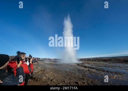 Reikiavik, Island. 21.. Okt. 2022. Während des Ausbruchs des Geysirs Strokkur im geothermischen Park Haukadalur werden Menschen gesehen, die den Geysir Strokkkur beobachten. Die geothermische Region in der Nähe des Flusses HvitÃ und der Stadt ReykjavÃ-k beherbergt den geothermischen Park Haukadalur. Hier befindet sich Strokkur, einer der berühmtesten Geysire Islands, der im Durchschnitt alle 4 bis 8 Minuten ausbricht, mit einer durchschnittlichen Höhe von 15 bis 20 Metern, manchmal bis zu 40 Meter. Im Park gibt es mehrere geothermische Merkmale wie Schlammbecken, Fumarolen, Algenablagerungen und andere Geysire daneben und um ihn herum. (Kreditbild: © Jorge Cas Stockfoto