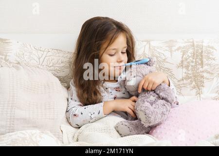 Lächelndes, selbstbewusstes, positives kleines Kind, Mädchen mit elektronischem Thermometer im Mund, umarmter Teddybär und beruhigendes Spielzeug im Bett. Covid 19, saisonal Stockfoto