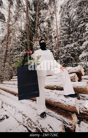 Ein Mädchen in einem weißen Anzug und Balaklava mit einem Paket Weihnachtsbäume im Winterwald an Silvester. Neujahrskonzept Stockfoto