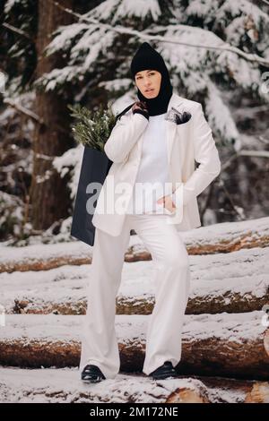 Ein Mädchen in einem weißen Anzug und Balaklava mit einem Paket Weihnachtsbäume im Winterwald an Silvester. Neujahrskonzept Stockfoto