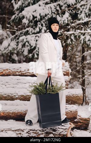 Ein Mädchen in einem weißen Anzug und Balaklava mit einem Paket Weihnachtsbäume im Winterwald an Silvester. Neujahrskonzept Stockfoto