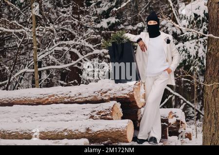 Ein Mädchen in einem weißen Anzug und Balaklava mit einem Paket Weihnachtsbäume im Winterwald an Silvester. Neujahrskonzept Stockfoto