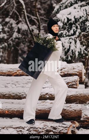 Ein Mädchen in einem weißen Anzug und Balaklava mit einem Paket Weihnachtsbäume im Winterwald an Silvester. Neujahrskonzept Stockfoto