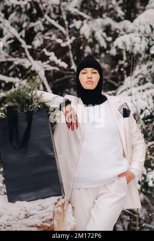 Ein Mädchen in einem weißen Anzug und Balaklava mit einem Paket Weihnachtsbäume im Winterwald an Silvester. Neujahrskonzept Stockfoto