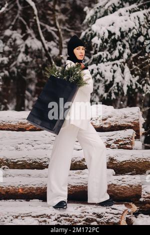 Ein Mädchen in einem weißen Anzug und Balaklava mit einem Paket Weihnachtsbäume im Winterwald an Silvester. Neujahrskonzept Stockfoto